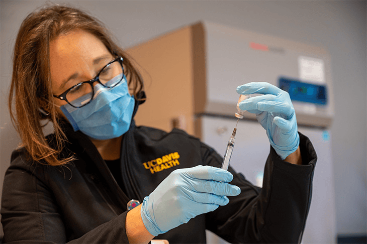 A woman in a lab coat and gloves holds a syringe, preparing for a medical procedure in a clinical setting.