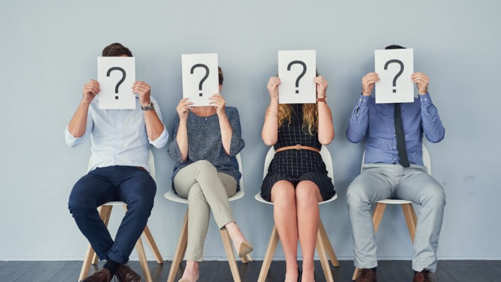 Four people sit in chairs can cover their faces with a paper with a question mark on it.